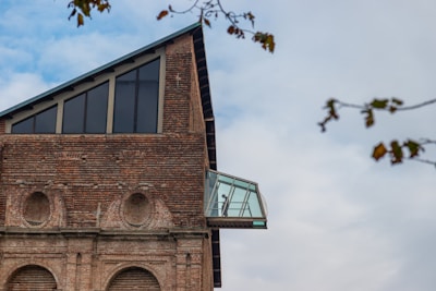 A brick building with a modern glass extension protruding from its upper floor. The extension features large windows and contains a silhouette of a person inside. The sky is partly cloudy, and some tree branches with sparse leaves are visible in the foreground.