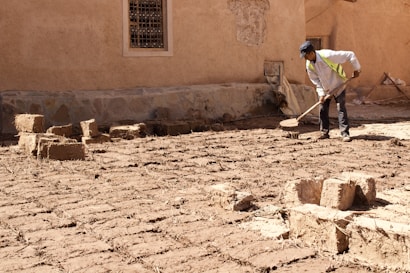 A person wearing a cap and reflective vest is using a manual tool to level or work on a surface made of adobe bricks. The setting features an earthen building wall with a window, giving it a rustic and traditional look.