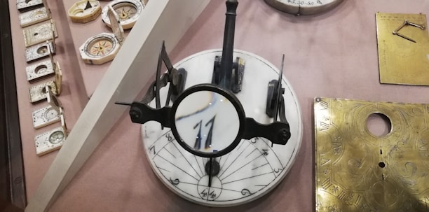 A display featuring various antique scientific instruments, including a sundial positioned centrally with a magnifying lens on top. Several ornate, compact compasses and dials are placed to the left. Two brass plates with engraved markings are on the right. The background is a soft beige, adding an antique feel to the arrangement.