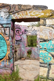 A partially destroyed stone structure adorned with colorful graffiti art. The vibrant designs include abstract patterns and cartoon-like characters, with old stone walls showing layers of graffiti accumulated over time. Vegetation is visible through the open windows and doorway, suggesting an abandoned and overgrown setting.