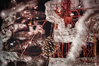 An intricate and festive holiday decoration featuring a combination of pinecones, star ornaments, and lace-like ribbons intertwined with red strings. The scene is lit up by small white lights, and fluffy white garlands add a wintry touch.