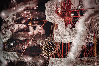 An intricate and festive holiday decoration featuring a combination of pinecones, star ornaments, and lace-like ribbons intertwined with red strings. The scene is lit up by small white lights, and fluffy white garlands add a wintry touch.