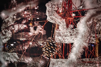 An intricate and festive holiday decoration featuring a combination of pinecones, star ornaments, and lace-like ribbons intertwined with red strings. The scene is lit up by small white lights, and fluffy white garlands add a wintry touch.
