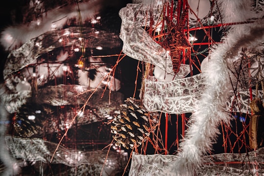 An intricate and festive holiday decoration featuring a combination of pinecones, star ornaments, and lace-like ribbons intertwined with red strings. The scene is lit up by small white lights, and fluffy white garlands add a wintry touch.