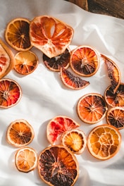 A variety of dried citrus slices are scattered on a white surface. The fruit slices display a range of vibrant colors, including deep reds, oranges, and yellows, with some pieces having a slightly desaturated tone. The textures of the dried fruit are highlighted by the sunlight, creating a natural and rustic aesthetic.