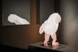 A geometric sculpture of a humanoid figure stands on a platform, crafted from sections of white paper or similar material. The figure has a simplified, abstract form with polygonal facets, giving it a digital or low-poly appearance. The setting is minimalistic, with a neutral-toned wall and floor. On the left side, a reflection of the sculpture appears in a mirror, reinforcing the symmetry of the scene.