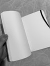 An open blank notebook held by a hand, with a black ribbon bookmark on the right page. The background features a textured surface with a circular pattern.