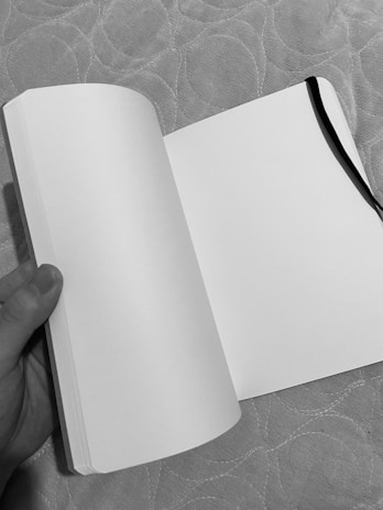 An open blank notebook held by a hand, with a black ribbon bookmark on the right page. The background features a textured surface with a circular pattern.