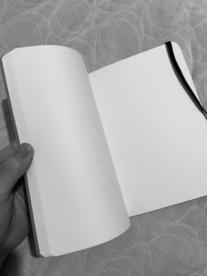 An open blank notebook held by a hand, with a black ribbon bookmark on the right page. The background features a textured surface with a circular pattern.