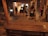 A dimly lit indoor setting with several people casually standing and walking about. The space appears to be a gallery or exhibition area, characterized by a wooden bench in the foreground, exposed beams, and a mixture of concrete and wooden textures. The warm ambient lighting and art pieces or installations can be seen through the door opening.