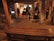 A dimly lit indoor setting with several people casually standing and walking about. The space appears to be a gallery or exhibition area, characterized by a wooden bench in the foreground, exposed beams, and a mixture of concrete and wooden textures. The warm ambient lighting and art pieces or installations can be seen through the door opening.