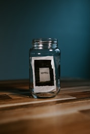 A glass jar sits on a wooden surface against a dark teal background. Inside the jar, there is a small paper or card with the word 'memories' written on it. The overall composition is simple, with a focus on the jar and the concept it represents.