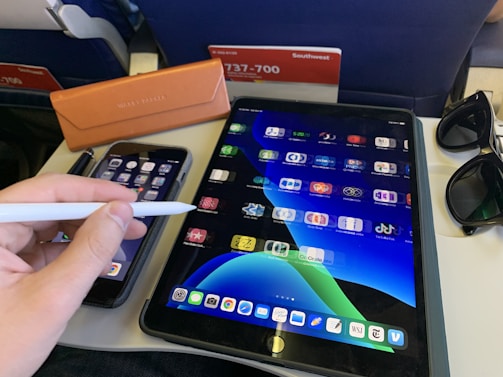 A person is holding a stylus near a tablet displaying various app icons. Next to the tablet, a smartphone is visible with its screen showing apps as well. There is a pair of sunglasses, an orange glasses case, and a pen on the table. The surroundings suggest an airplane setting, with the back of a seat and a safety information card in view.