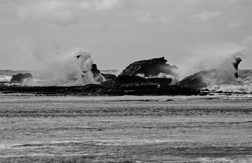 Foto en escala de grises del hombre parado en una formación rocosa cerca del mar