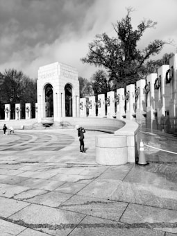 A black and white photograph of a large stone monument labeled 'Atlantic' with an archway and a series of columns, each adorned with wreaths. The scene includes a person taking photographs and another couple walking. The sky is partly cloudy, and tall trees can be seen in the background.