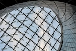 A futuristic architectural design featuring a large curved glass structure, with metal supports forming a geometric pattern against a backdrop of blue sky and clouds.