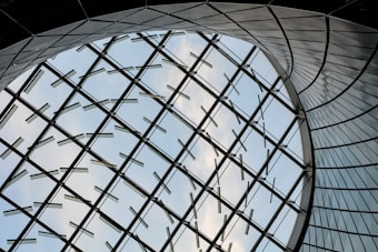 A futuristic architectural design featuring a large curved glass structure, with metal supports forming a geometric pattern against a backdrop of blue sky and clouds.