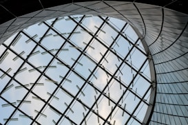 A futuristic architectural design featuring a large curved glass structure, with metal supports forming a geometric pattern against a backdrop of blue sky and clouds.