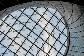 A futuristic architectural design featuring a large curved glass structure, with metal supports forming a geometric pattern against a backdrop of blue sky and clouds.