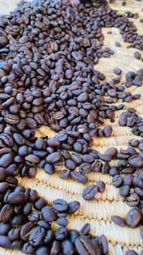 A close-up view of roasted coffee beans scattered across a woven straw mat, providing an earthy and textured background. The beans are rich and dark in color, suggesting a deep roast.