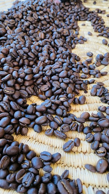 A close-up view of roasted coffee beans scattered across a woven straw mat, providing an earthy and textured background. The beans are rich and dark in color, suggesting a deep roast.