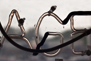 A close-up view of a neon sign with a portion of the letters 'AGE' visible. The neon tubes are unlit, with wiring and connectors attached. The background appears blurred, possibly showing an outdoor scene.