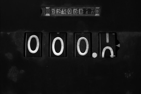An old mechanical counter with multiple rotating wheels displaying various digits. The main section of the counter shows the numbers 0000 prominently, while a smaller section at the top shows the number 0250084. The device appears worn and is set against a dark background.