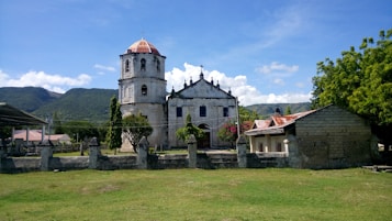 A historic stone church stands prominently with a distinctive bell tower, surrounded by a low stone wall and lush greenery. The setting includes vibrant trees and a backdrop of green hills under a clear blue sky. Adjacent structures feature weathered, rusted roofs, adding to the rustic charm of the scene.