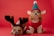 Two small dogs dressed in festive holiday costumes sit against a red background. The dog on the left wears a reindeer costume with antlers, while the dog on the right is dressed as an elf with a green hat and plaid trim, featuring elf-like ears.