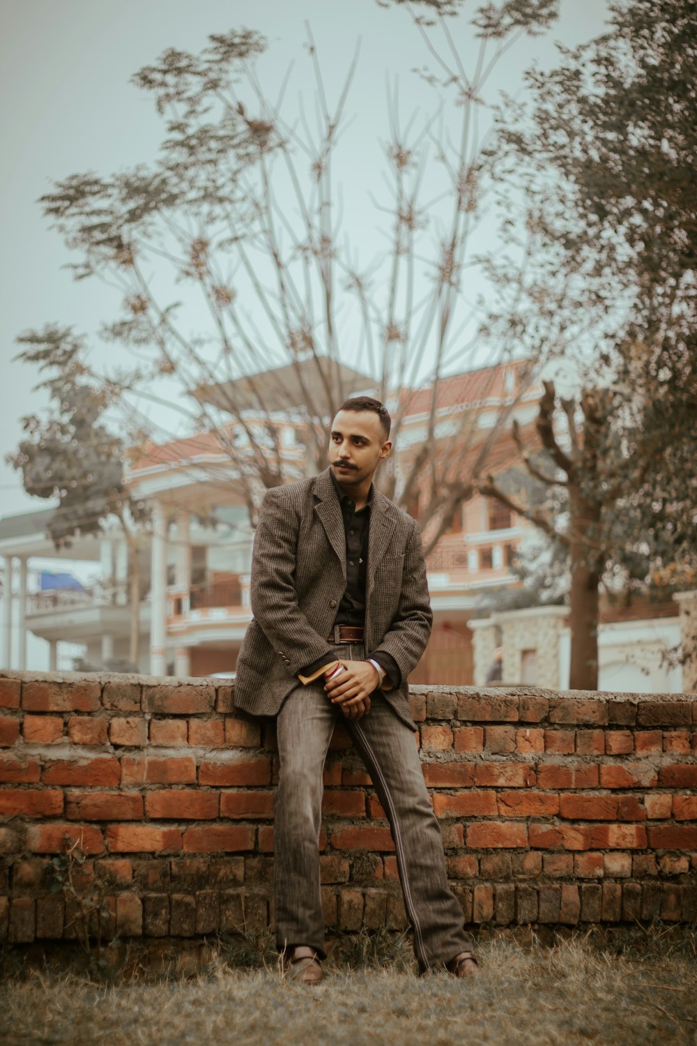 Mann im grauen Mantel sitzt tagsüber auf brauner Ziegelmauer