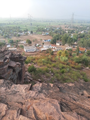 A rural landscape featuring rocky terrain in the foreground, leading to a small village with scattered houses and greenery. The scene includes fields and trees stretching into the distance, with power lines and towers visible against a hazy sky.