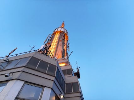 A tall, metallic tower structure with illuminated orange lights along its vertical edges, against a clear blue sky. The base part of the structure includes several glass and metal panels with a few visible security cameras.