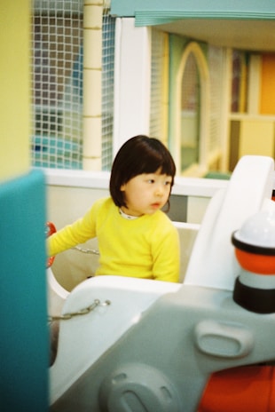 A young child wearing a bright yellow sweater is seated in a colorful indoor play area. The child appears to be sitting on a toy vehicle with a steering wheel, surrounded by pastel-colored walls and safety netting.