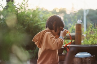 A young child with short hair is holding a red and black camera, appearing to be focused on taking a photo. The child is wearing a light brown hoodie and is surrounded by lush greenery. The background appears blurred, with soft natural lighting indicating a possibly early morning or late afternoon setting.