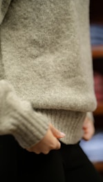 A close-up of a person wearing a light gray woolen sweater, with hands adjusting the bottom hem. The focus is on the texture of the sweater and the neutral colors.