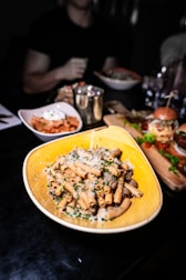 A plate of pasta topped with grated cheese and herbs is prominently displayed on a dark table. Surrounding it are other dishes like a bowl of salad, a small bowl with a red sauce, and a burger on a wooden platter. The background shows blurred figures and additional dining items.