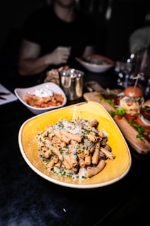 A plate of pasta topped with grated cheese and herbs is prominently displayed on a dark table. Surrounding it are other dishes like a bowl of salad, a small bowl with a red sauce, and a burger on a wooden platter. The background shows blurred figures and additional dining items.