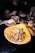 A plate of pasta topped with grated cheese and herbs is prominently displayed on a dark table. Surrounding it are other dishes like a bowl of salad, a small bowl with a red sauce, and a burger on a wooden platter. The background shows blurred figures and additional dining items.