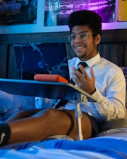 A person wearing glasses and a white shirt with a black tie is sitting on a bed, using a laptop placed on a folding bed table. The person is smiling and appears to be engaged in a video call or online meeting. The room has dim lighting with a blue and purple hue, and there are posters on the wall in the background.