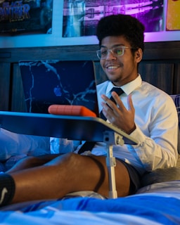 A person wearing glasses and a white shirt with a black tie is sitting on a bed, using a laptop placed on a folding bed table. The person is smiling and appears to be engaged in a video call or online meeting. The room has dim lighting with a blue and purple hue, and there are posters on the wall in the background.