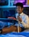 A person wearing glasses and a white shirt with a black tie is sitting on a bed, using a laptop placed on a folding bed table. The person is smiling and appears to be engaged in a video call or online meeting. The room has dim lighting with a blue and purple hue, and there are posters on the wall in the background.
