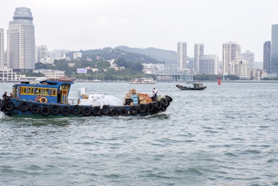 A small cargo boat is sailing in a wide river or coastal area with a city skyline in the background. The boat is carrying various goods, including packages and bags, with large tires used as fenders along its sides. The skyline features several high-rise buildings, hills in the distance, and cloudy weather.