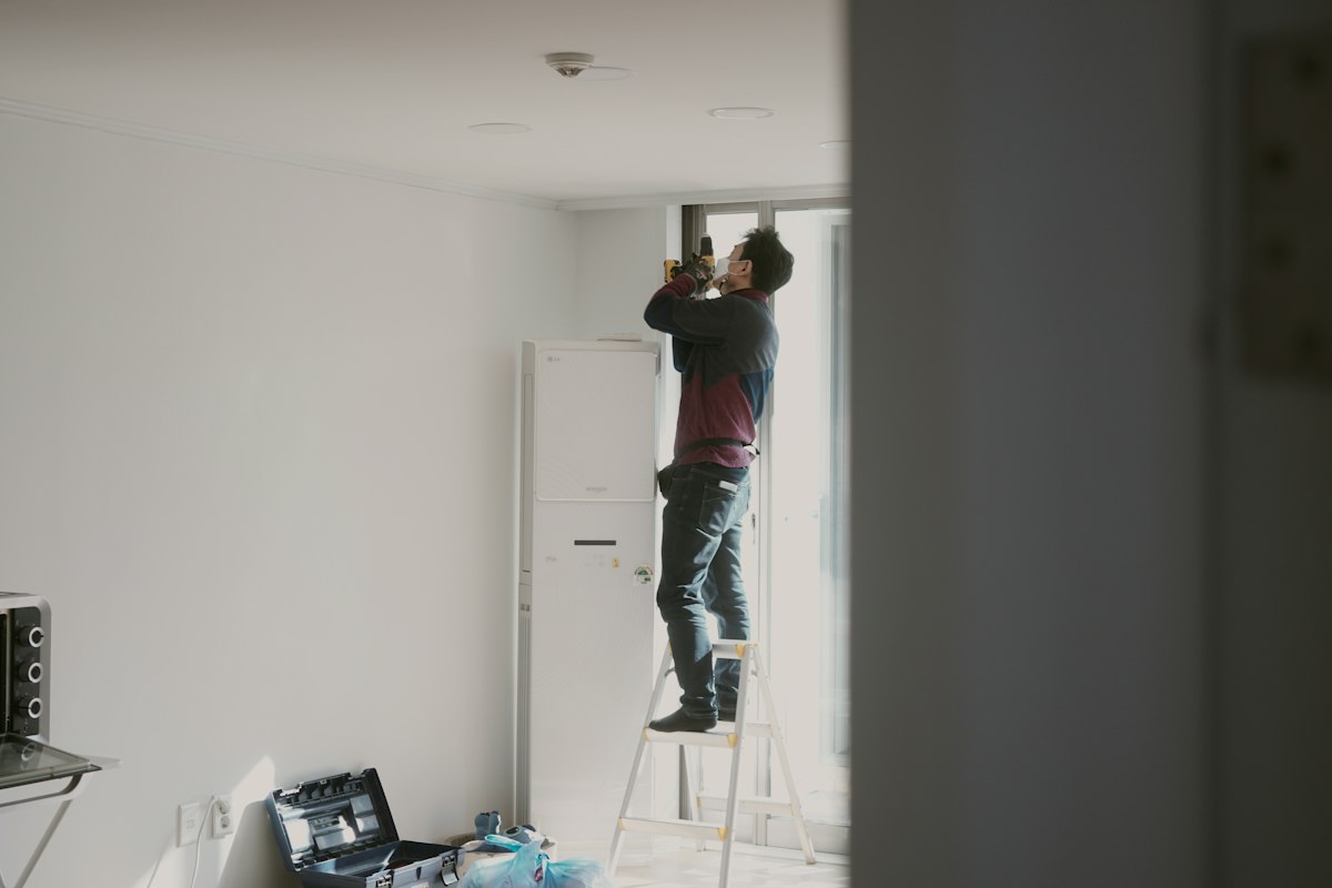 A handyman in a face mask stands on a stepladder in a bright room, using a drill near a window, with a toolbox on the floor