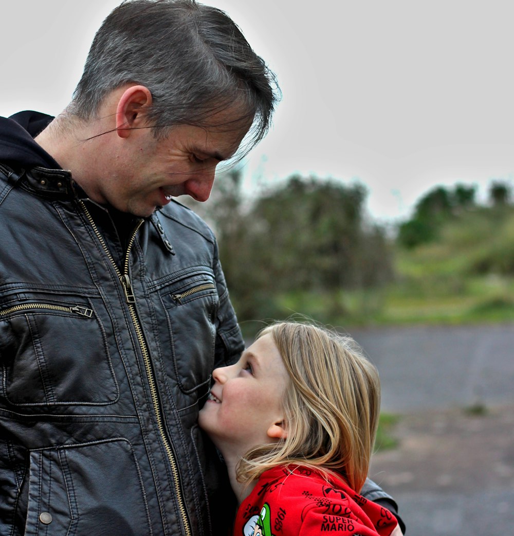 man in black leather jacket kissing woman in red hoodie during daytime