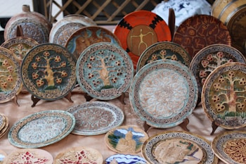 A collection of intricately designed ceramic plates is displayed on a table. Each plate features unique patterns and motifs, including depictions of trees, geometric designs, and cultural symbols. The decorations on the plates are detailed, highlighting artistic craftsmanship. Some plates are mounted on stands for better visibility.
