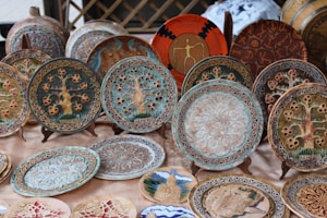 A collection of intricately designed ceramic plates is displayed on a table. Each plate features unique patterns and motifs, including depictions of trees, geometric designs, and cultural symbols. The decorations on the plates are detailed, highlighting artistic craftsmanship. Some plates are mounted on stands for better visibility.