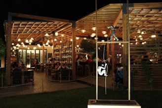 An outdoor restaurant with wooden structures and warm lighting creates a cozy and inviting atmosphere. Patrons are seated at tables, engaged in conversation and dining. Shelves filled with wine bottles line the walls, enhancing the ambiance. Sculptures of climbing figures are visible in the foreground, adding artistic interest.