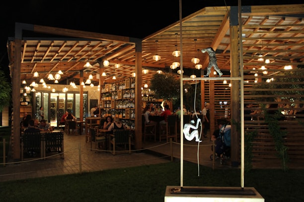 An outdoor restaurant with wooden structures and warm lighting creates a cozy and inviting atmosphere. Patrons are seated at tables, engaged in conversation and dining. Shelves filled with wine bottles line the walls, enhancing the ambiance. Sculptures of climbing figures are visible in the foreground, adding artistic interest.