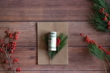 A rolled-up US dollar bill is tied with a red and white string, placed on a brown card. Pine branches with red berries surround the card on a wooden surface.
