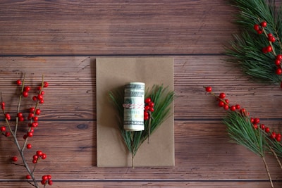 A rolled-up US dollar bill is tied with a red and white string, placed on a brown card. Pine branches with red berries surround the card on a wooden surface.